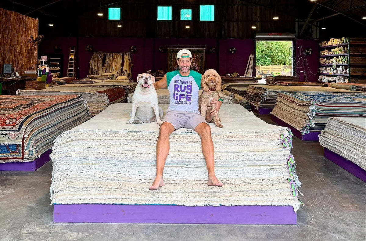 Banana Manor owner sitting on a stack of whitewashed rugs with the two shop dogs